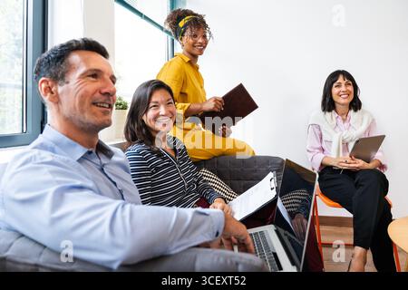 Quatre collègues souriants collaborent sur un projet, en utilisant un ordinateur portable, une tablette et des documents, démontrant le travail d'équipe et l'innovation dans un bureau moderne Banque D'Images