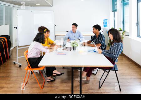 Cinq hommes d'affaires sont assis autour d'une table blanche dans un bureau moderne, collaborant à un projet utilisant des ordinateurs portables et des tablettes, discutant des stratégies Banque D'Images