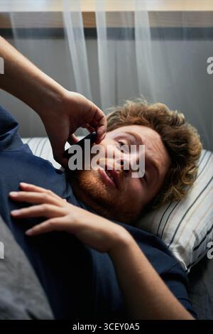 Gros plan vertical moyen d'un jeune homme caucasien se relaxant au lit le matin parlant au téléphone avec sa petite amie Banque D'Images