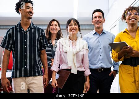 Groupe de gens d'affaires multiethniques marchant ensemble dans le couloir du bureau, souriant et tenant des dossiers et une tablette, profitant de leur travail dans un positi Banque D'Images