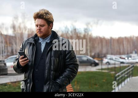 Portrait horizontal moyen de jeune homme caucasien portant une tenue décontractée élégante marchant à l'extérieur le jour d'automne regardant quelque chose sur smartphone Banque D'Images