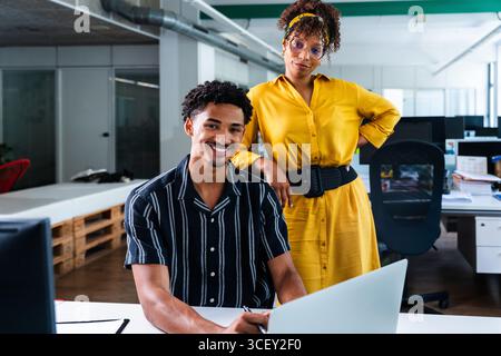 Deux jeunes collègues souriants, un homme et une femme, travaillent ensemble sur un ordinateur portable dans un bureau moderne de start-up, montrant le travail d'équipe et collaborati Banque D'Images