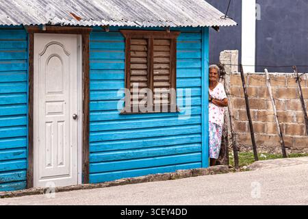 Cueva de chico, république dominicaine 20 août 2025 : femme dominicaine âgée debout près de sa vibrante maison bleue à cueva de chico, reflétant la vie locale Banque D'Images