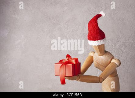 Homme en bois dans un chapeau de père noël tient un cadeau dans une boîte rouge Banque D'Images