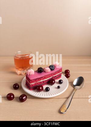 Une tranche de gâteau en couches avec glaçage rose, garnie de bleuets et de cerises, servi sur une assiette blanche. Accompagné d'un verre de thé et d'une déco Banque D'Images