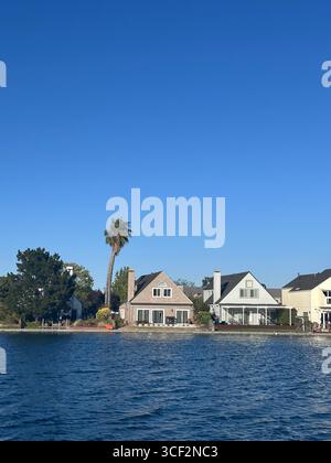 Une rangée de maisons au bord de l'eau avec un grand palmier et un ciel bleu clair, vu de l'autre côté de l'eau par une journée ensoleillée brillante. Banque D'Images