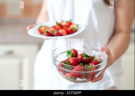 Gros plan des mains féminines tenant deux assiettes avec des fraises entières ​​and tranchées, préparant une collation saine Banque D'Images