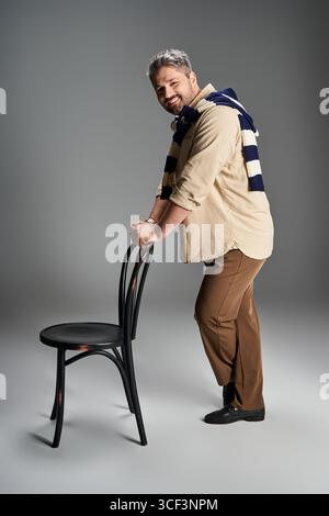 Un bel homme pose avec confiance dans une tenue chic, tenant sur une chaise noire dans un studio élégant. Banque D'Images