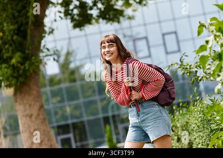 Jeune femme en pull rayé et short marche joyeusement à travers le campus, immergée dans ses études. Banque D'Images