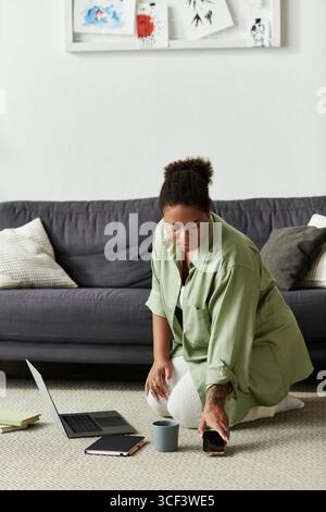 Une femme afro-américaine s'agenouille à la maison, équilibrant confort décontracté et concentration au travail. Banque D'Images