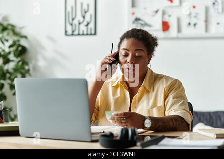 Une femme afro-américaine travaille à distance alors qu'elle est assise à son bureau dans son appartement. Banque D'Images