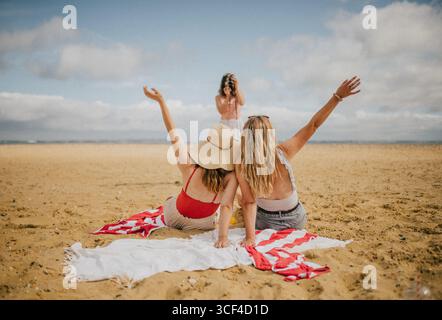 Deux femmes sont assises sur une plage, l'une d'elles tenant un appareil photo. L'autre femme porte un haut rouge et un chapeau blanc. La scène est détendue et insouciante Banque D'Images