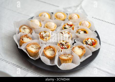 Un délicieux assortiment de mini-hors-d'œuvre magnifiquement disposés sur un plateau rond foncé. chaque friandise en bouchée est enveloppée dans une tasse en papier blanc, showcas Banque D'Images