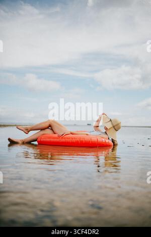 Une femme est allongée sur un radeau gonflable rouge dans l'océan. Elle porte un chapeau de paille et lit un livre. La scène est paisible et relaxant, avec le w Banque D'Images