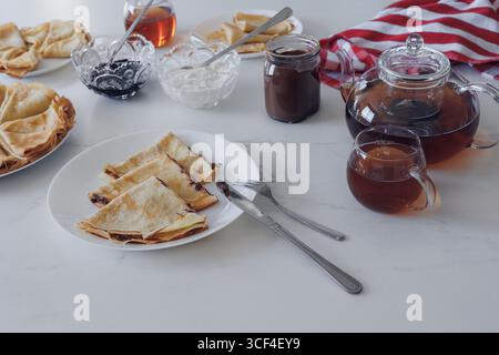 Crêpes traditionnelles avec confiture, crème sure et thé servis sur table blanche. Vue de dessus, ambiance maison confortable et concept culinaire Banque D'Images