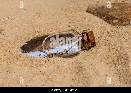 Bouteille en verre avec bouchon en liège contenant du papier enroulé avec coordonnées manuscrites, à moitié enfouie dans le sable de plage Banque D'Images