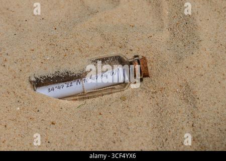 Bouteille en verre avec un message enroulé montrant des coordonnées géographiques se trouvant sur le sable d'une plage Banque D'Images