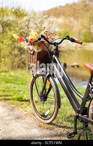 Un vélo avec un panier rempli de fleurs colorées se dresse sur un chemin dans un paysage printanier idyllique. Tour de vélo ensoleillé en toile de fond pour le tourisme, les loisirs et le style de vie respectueux de l'environnement. Espace pour le texte. Banque D'Images