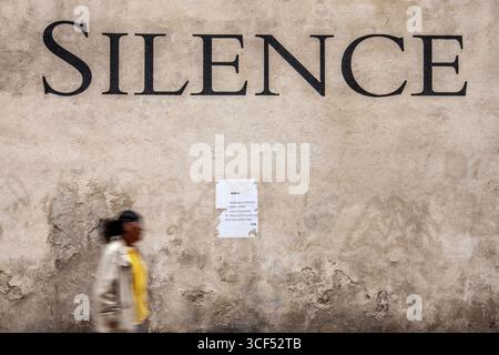 Grand lettrage 'silence' sur un mur beige avec une personne passant devant dans un environnement urbain Banque D'Images