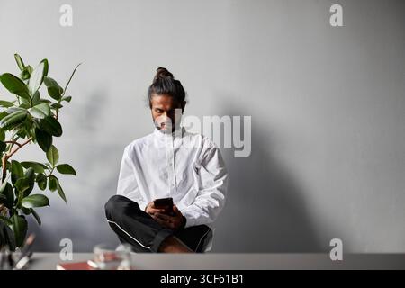 Portrait horizontal moyen studio de jeune homme indien portant chemise blanche élégante assis détendu dans la salle de bureau surfant sur Internet sur smartphone Banque D'Images