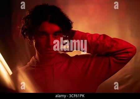 Portrait horizontal moyen en gros plan d'un beau jeune homme aux cheveux ondulés portant un sweat à capuche debout dans une lumière rouge et orange posant sur l'appareil photo Banque D'Images