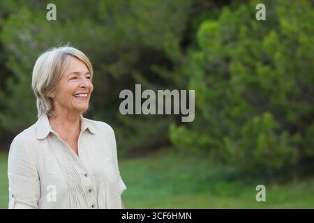 Femme senior debout souriant dans un champ herbeux par des arbres à feuilles persistantes portant un chemisier en lin, espace de copie Banque D'Images