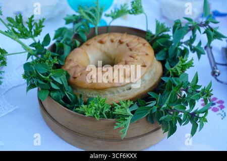 Un koulouri doux crétois traditionnel, un pain en forme d'anneau, est élégamment exposé dans un cadre rustique en forme de tamis, entouré d'herbes fraîches. Banque D'Images