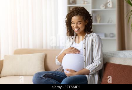 Femme afro-américaine enceinte souriante assise sur un canapé, touchant le ventre avec amour Banque D'Images