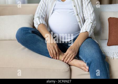Femme afro-américaine enceinte assise sur un canapé, tenant la cheville gonflée avec les mains Banque D'Images