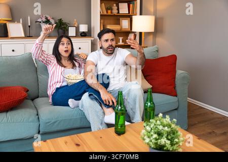 Jeune homme de couple et femme assis à la maison sur le canapé prenant une pause après une journée de travail stressante regarder un jeu de sport ou un film à la télévision, manger du pop-corn et du drin Banque D'Images