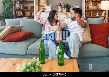 Jeune homme de couple et femme assis à la maison sur le canapé prenant une pause après une journée de travail stressante regarder un jeu de sport ou un film à la télévision, manger du pop-corn et du drin Banque D'Images