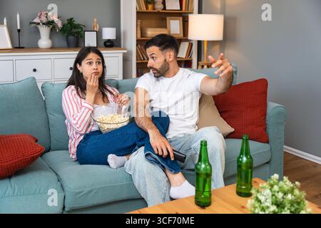 Jeune homme de couple et femme assis à la maison sur le canapé prenant une pause après une journée de travail stressante regarder un jeu de sport ou un film à la télévision, manger du pop-corn et du drin Banque D'Images