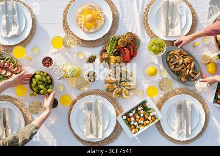 Vue aérienne d'un groupe multiethnique de jeunes adultes et mains d'âge moyen atteignant pour des hors-d'œuvre assortis, salades, fruits et boissons sur une table en plein air ensemble pour un repas partagé Banque D'Images