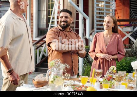 Groupe d'homme caucasien d'âge moyen, jeune femme caucasienne et homme du moyen-Orient avec la barbe debout à l'extérieur autour de la table avec de la nourriture et des boissons, souriant et conversant Banque D'Images