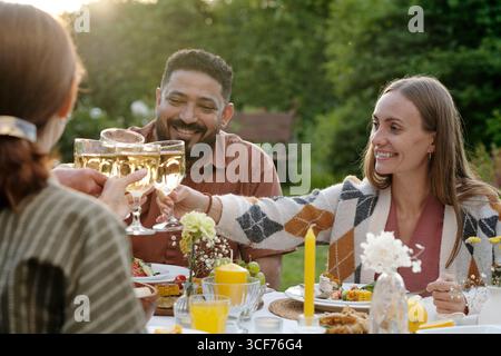 Groupe de membres de la famille assis à l'extérieur à prendre des verres de vin pendant la cueillette, souriant et appréciant le repas ensemble à table Banque D'Images