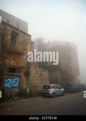 Vue sur la rue brumeuse avec d'anciens bâtiments en pierre couverts de brouillard du matin et des voitures garées le long de la route étroite dans l'historique Enna Sicile Italie Banque D'Images