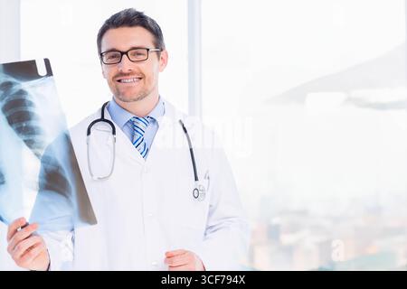 Médecin masculin examinant un film radiographique thoracique à la fenêtre de la clinique portant une blouse de laboratoire et un stéthoscope Banque D'Images