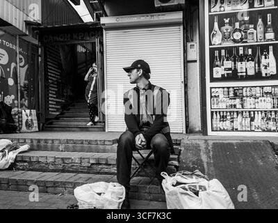 Kiev, Ukraine - 20 juillet 2025 : vendeur de rue assis sur une chaise pliante devant son stalle avec des sacs de marchandises à ses pieds, tandis qu'une femme monte les escaliers dans un marché aux puces ukrainien Banque D'Images