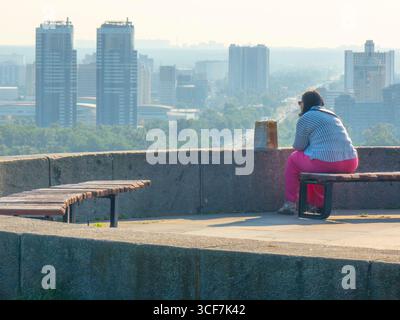 Kiev, Ukraine - 20 juillet 2025 : femme assise sur un banc contemplant le paysage urbain de Kiev depuis le point de vue du Parc de la gloire éternelle, profitant d'un panorama urbain avec gratte-ciel et trafic Banque D'Images