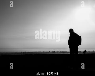 Kiev, Ukraine - 20 juillet 2025 : silhouette noire et blanche d'un homme portant un sac à dos regardant le paysage urbain de Kiev depuis le Parc de la gloire éternelle, Ukraine, avec un pigeon à proximité Banque D'Images
