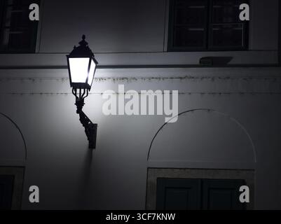 Un lampadaire à l'ancienne attaché à un mur de bâtiment blanc, brillant brillamment dans la nuit sombre. Banque D'Images