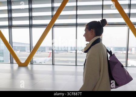 Un voyageur se promène dans un terminal d'aéroport moderne, transportant un sac à dos. De grandes fenêtres présentent des avions sur le tarmac et un voyage animé Banque D'Images