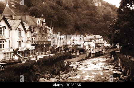 Cette vue aux tons sépia depuis East Lyn Bridges à Lynmouth capture un mélange harmonieux d'architecture et de nature. Le Royal Hotel et le Riverside Café sont nichés au bord de la rivière, leurs toits abrupts et cheminées faisant écho au design européen traditionnel. Des murs de pierre guident l'eau qui coule, tandis que des arbres et une colline boisée s'élèvent en arrière-plan. La scène évoque un sentiment de charme tranquille et de continuité historique, où l'environnement bâti complète le terrain accidenté de la vie du village côtier du Devon. Banque D'Images