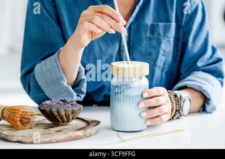 Femme boit Blue Iced Matcha Latte dans la cuisine près des accessoires pour sa préparation Banque D'Images
