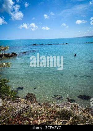Des eaux cristallines se baignant doucement contre la rive rocheuse sous un ciel bleu vif avec des nuages blancs moelleux, créant une scène sereine et idyllique Banque D'Images