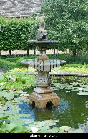 Une charmante fontaine en pierre verse de l'eau dans un étang tranquille rempli de nénuphars vibrants et entouré de riches plantes vertes. Banque D'Images