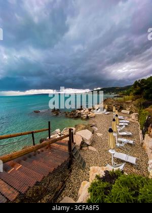 Chaises longues altérées, parasols pliés reposant sur la côte rocheuse sous un ciel nuageux sombre à Rocca San Giovanni, Abruzzes Banque D'Images