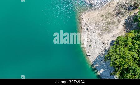 Les eaux turquoises se tapissent doucement contre un rivage rocheux, entouré d'arbres verdoyants. La lumière du soleil scintille sur la surface, créant un calme et un calme Banque D'Images