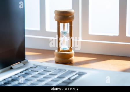 Gros plan d'un petit sablier à côté d'un ordinateur portable. Productivité, délai, concept de gestion du temps. Banque D'Images