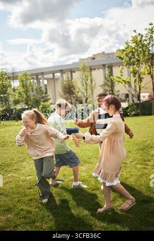Groupe d'enfants multiethniques jouant avec la machine à bulles sur l'herbe, riant et courant à l'extérieur, deux filles et deux garçons interagissant énergiquement dans le cadre du parc Banque D'Images